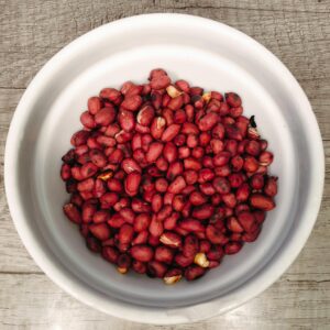 A white bowl filled with roasted red peanuts, viewed from above on a wooden surface.