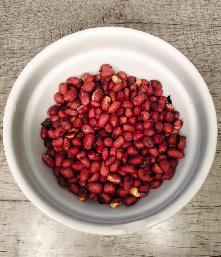 A white bowl filled with roasted red peanuts, viewed from above on a wooden surface.